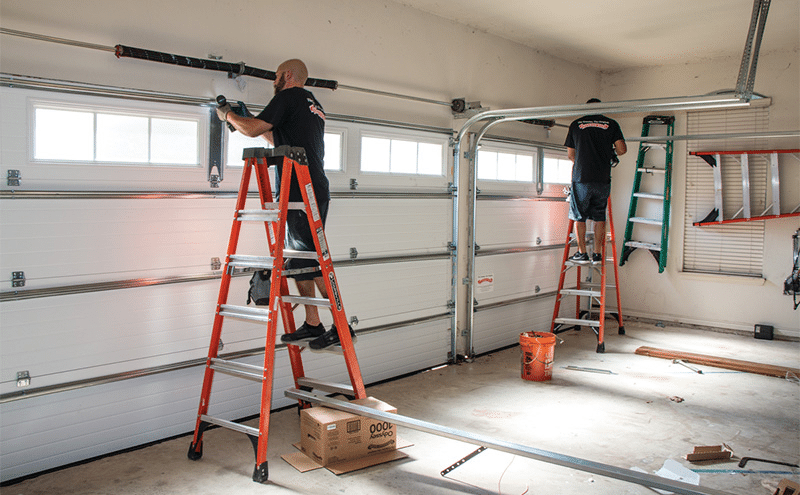 Two garage door technicians install and service white residential garage doors inside a clean two-car garage using orange ladders and professional tools.