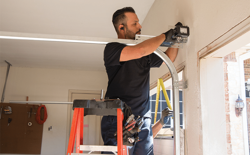 Garage door technician installs a residential garage door track and hardware while standing on a ladder inside a home garage.